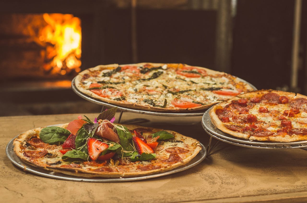 menu-03 Trio of gourmet pizzas with unique toppings near a wood-fired oven, ideal for food enthusiasts.